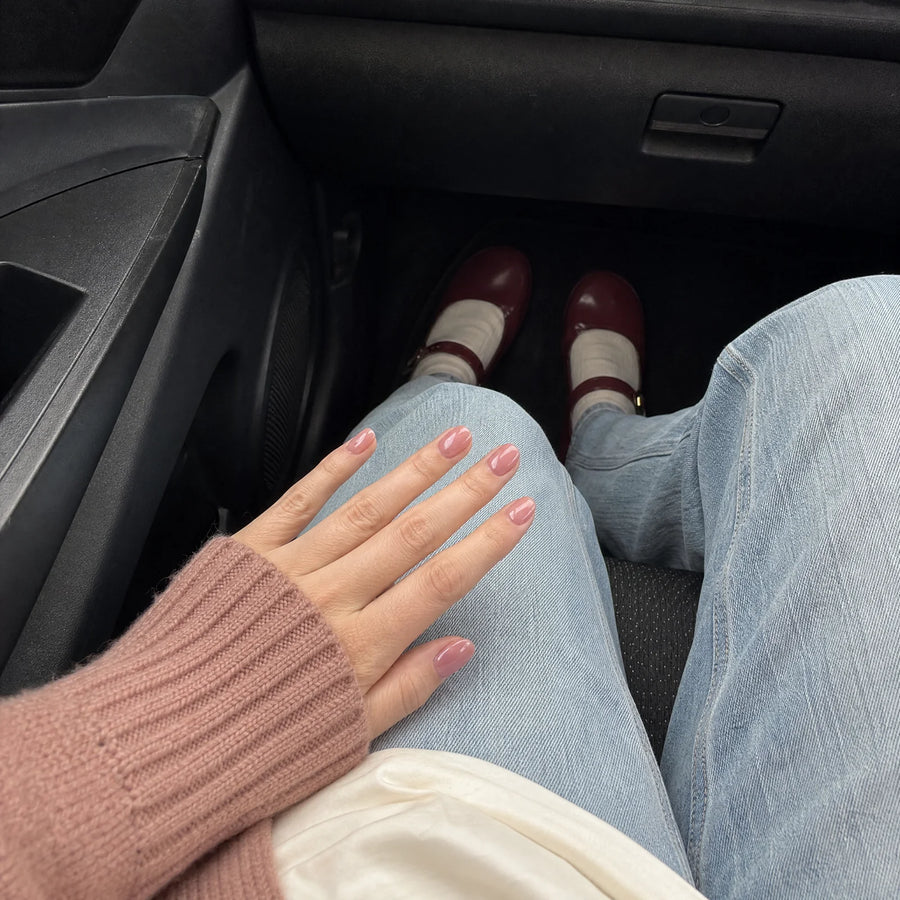 Face-free car-view campaign photo showing a dusty-rose manicure over light-wash jeans with burgundy shoes visible.